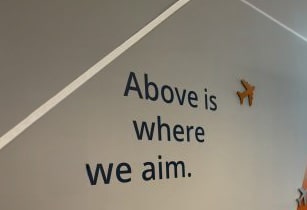 Schriftzug "Above is where we aim" mit Flugzeug-Icon an der Wand.