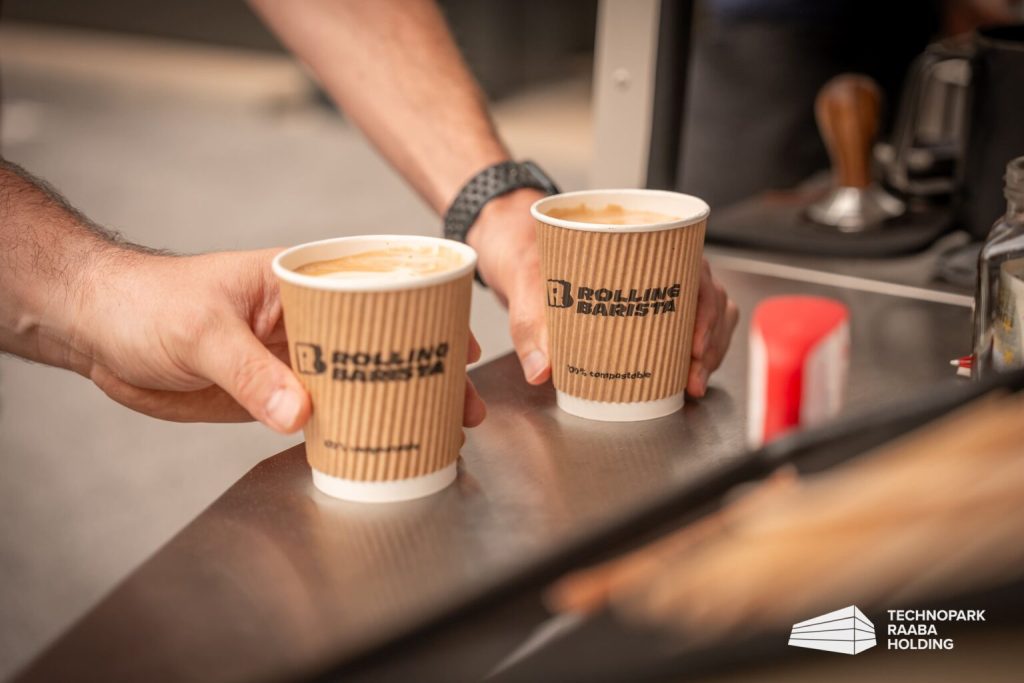 Zwei Hände reichen zwei Pappbecher mit Rolling Barista Logo über eine Theke.