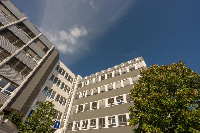 Bürogebäude des Technopark Raaba vor blauem Himmel, umgeben von grünen Bäumen.