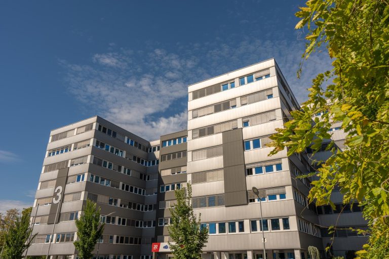 Mehrstöckiges Bürogebäude TP 3 bei sonnigem Wetter, umgeben von Bäumen.