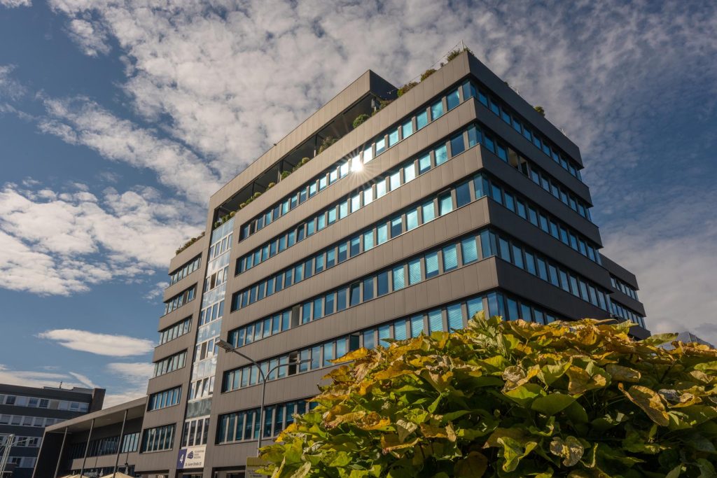 Moderner Bürokomplex unter blauem Himmel mit Wolken, im Vordergrund grüne Pflanzen.
