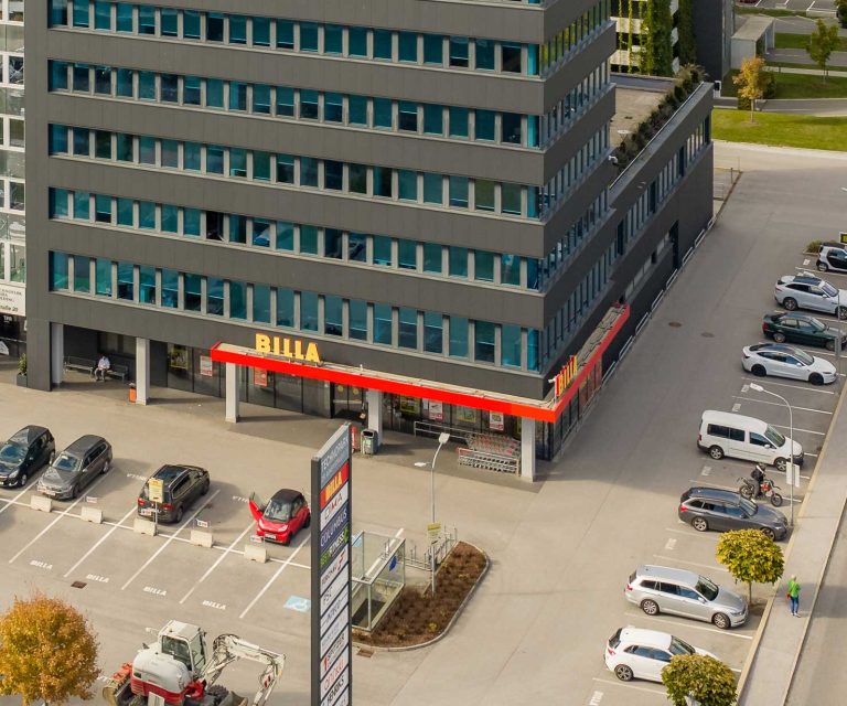 Parkplatz vor modernem Bürogebäude mit Billa-Supermarkt, mehrere geparkte Autos und grüne Bäume, Ansicht von oben.