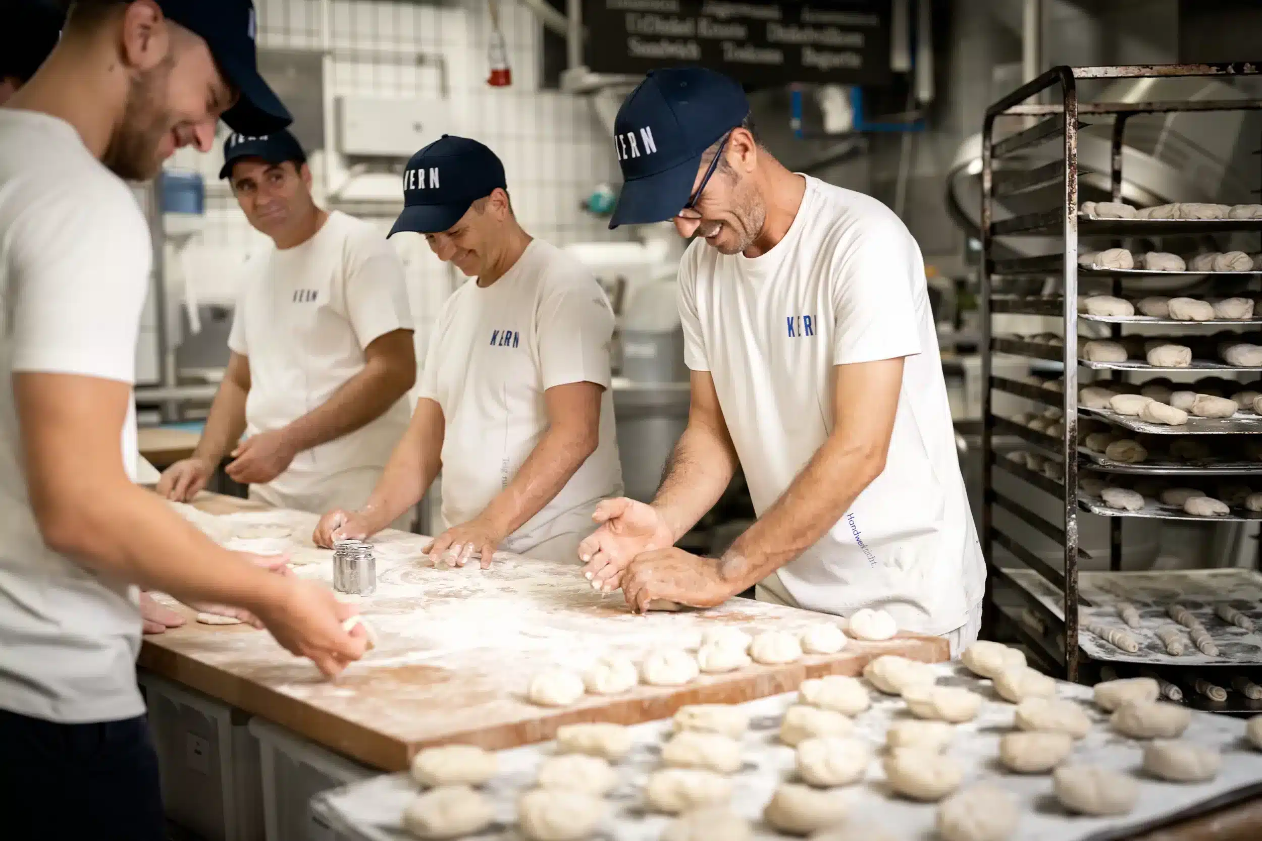 Mitarbeiter kneten Teig in moderner Bäckerei, Lächeln und Teamarbeit deutlich sichtbar.