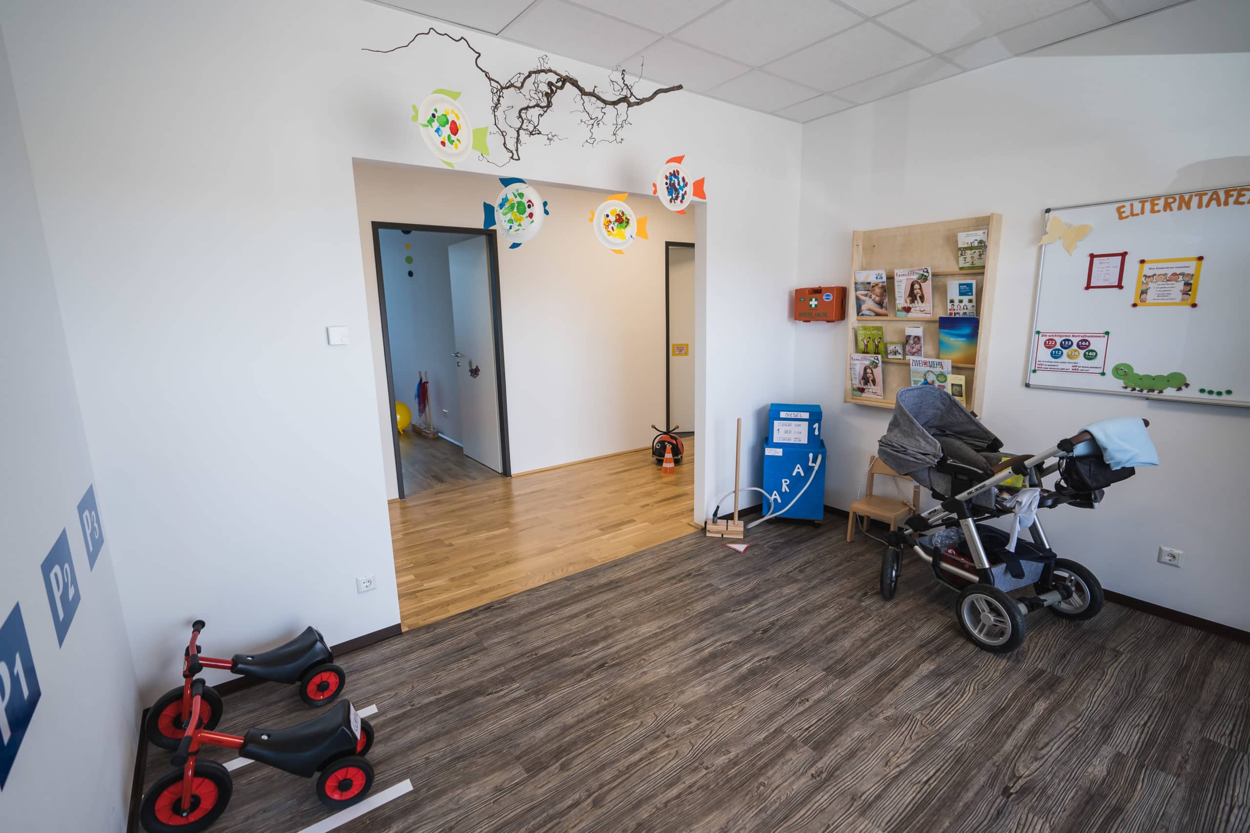 Fahrräder und Kinderwagen in einem hellen Raum mit Holzboden und farbigen Leuchten an der Decke.