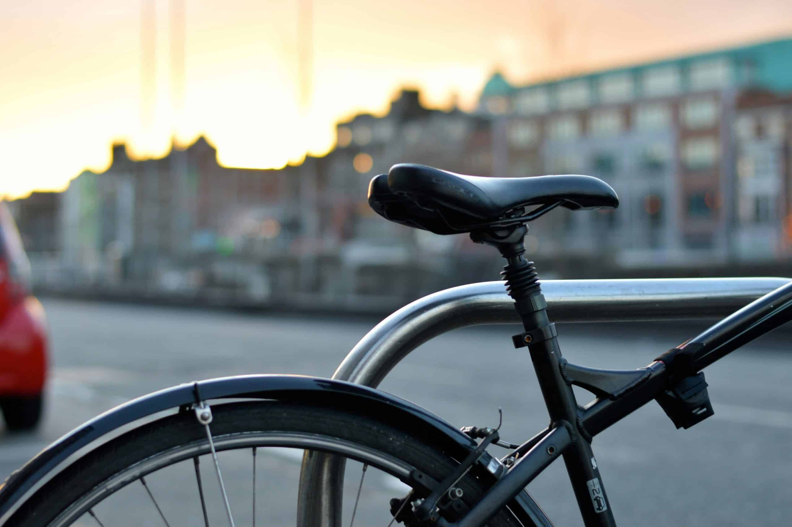 Fahrrad an einem Außenständer bei Sonnenuntergang; unscharfe Gebäude im Hintergrund.