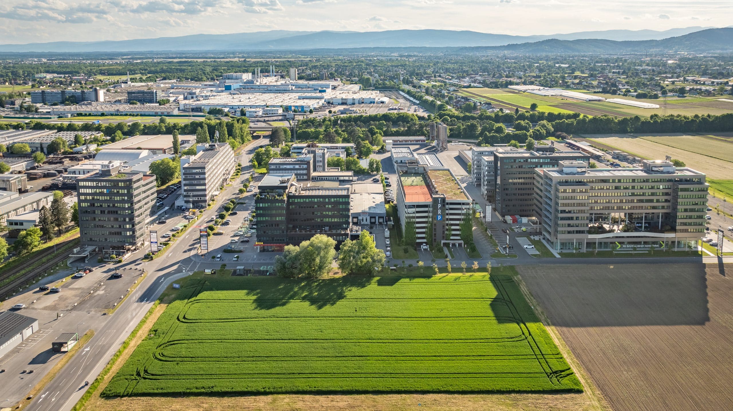 Luftaufnahme des Technopark Raaba umgeben von Feldern und Straßen, Stadt und Berge im Hintergrund.