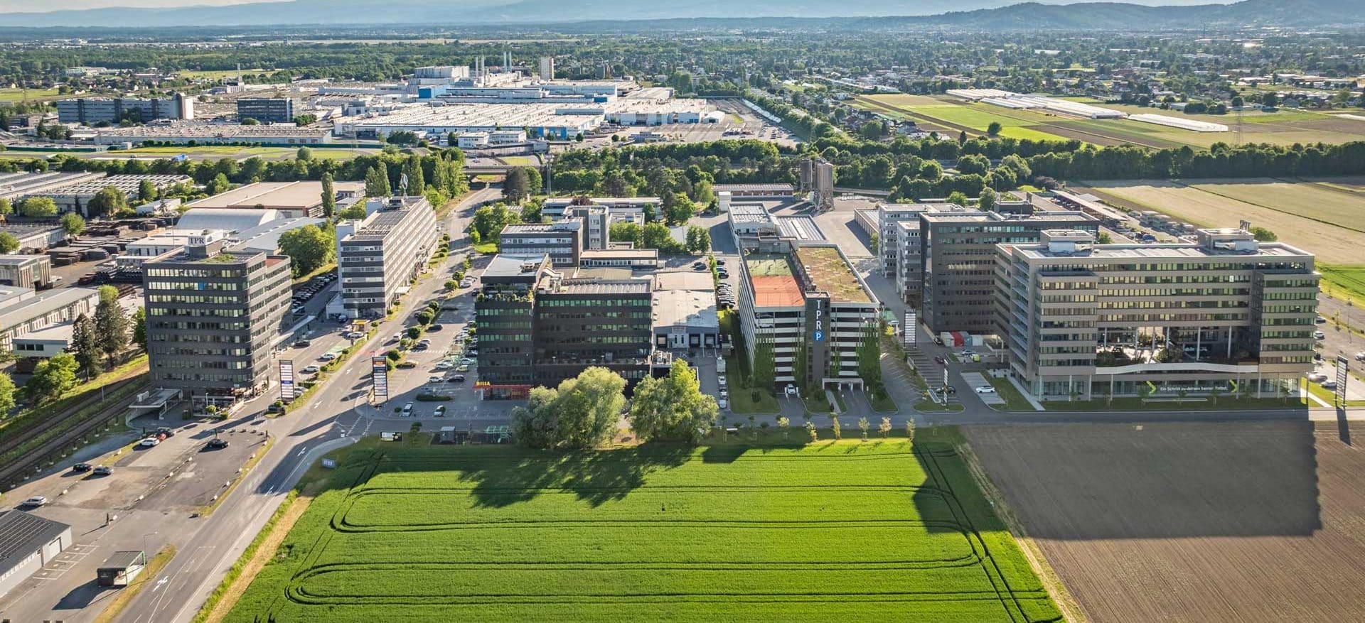 Luftaufnahme des Technopark Raaba mit grünen Feldern und Gebäuden vor einem Bergpanorama.